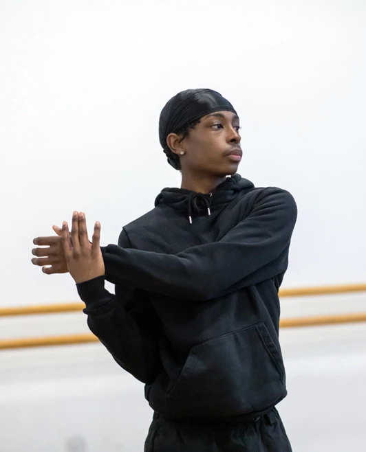 Male student wearing a black hoodie, posing with arms stretched in a dance studio, showing focused concentration during rehearsal. Male student wearing a black hoodie, posing with arms stretched in a dance studio, showing focused concentration during rehearsal.